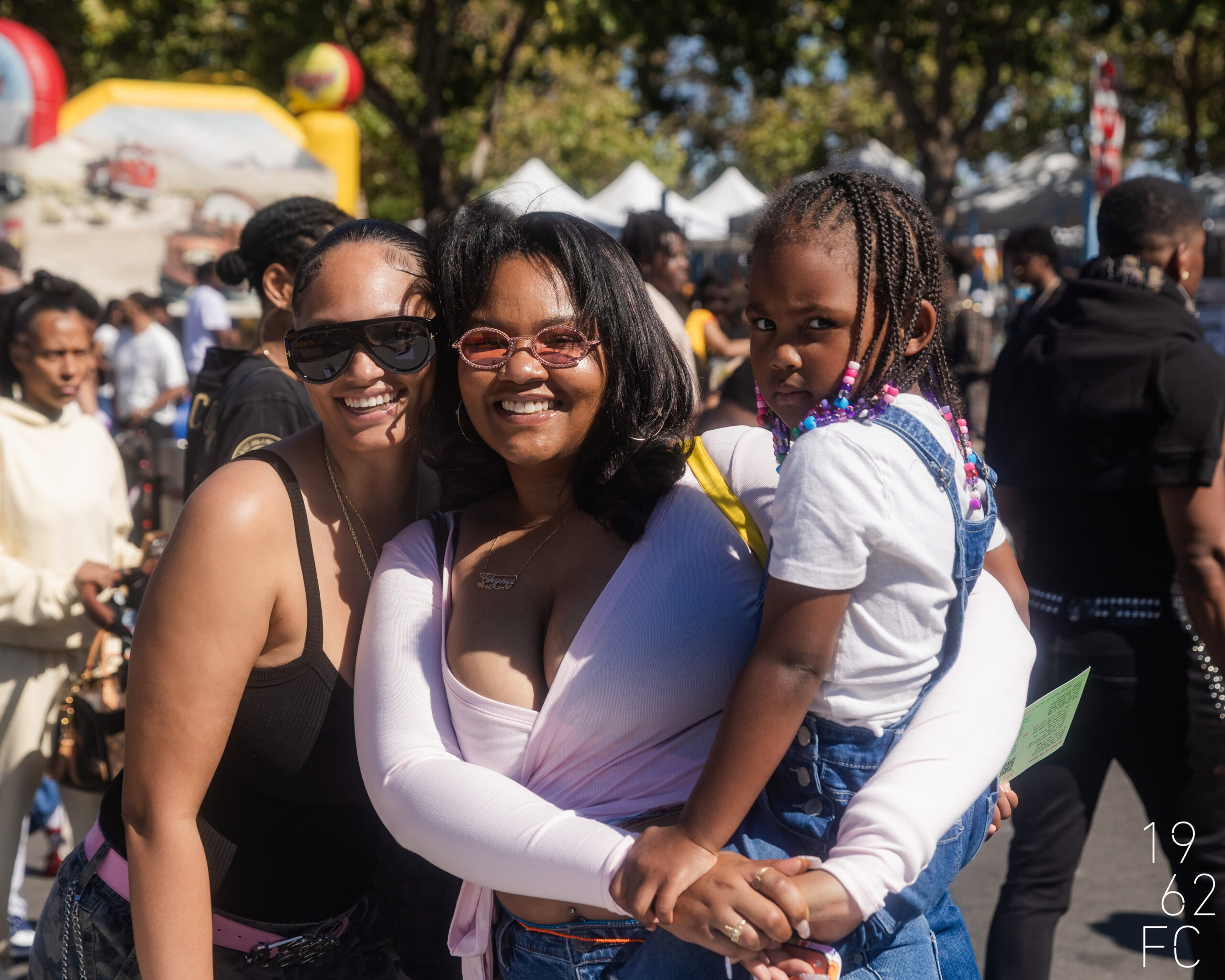 Group of smiling women and a girl enjoying outdoor festival with tents and trees.
