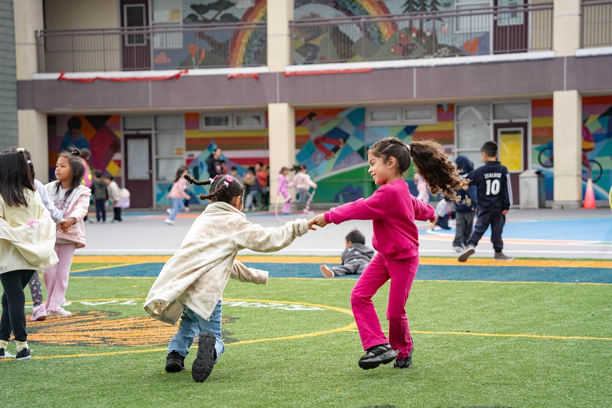 San Francisco School Fields 2026: Monroe Elementary Two young girls hold hands and spin on a colorful turf field at a San Francisco elementary school.